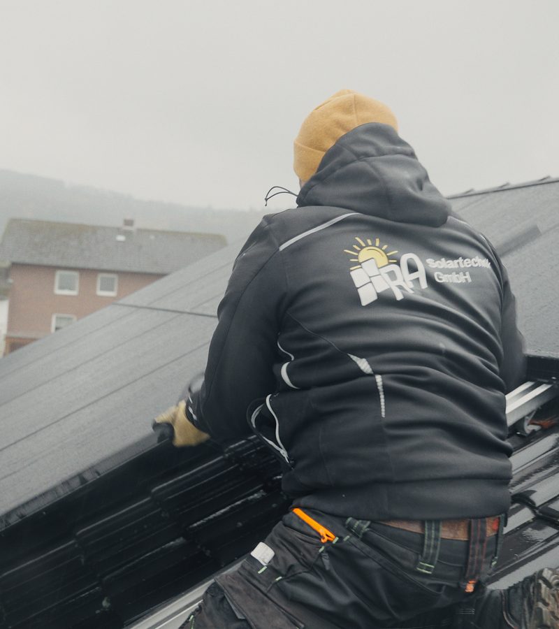 Monteur von RA Solartechnik mit Firmenlogo auf der Jacke verlegt schwarze Solarmodule auf einem Ziegeldach