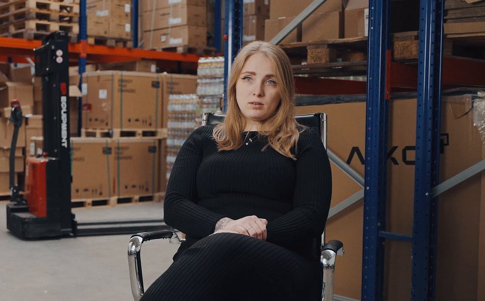 Michelle-Kathrin Börkei von RA Solartechnik sitzt im Lager vor Regalen mit Paletten und Kartons, rechts Modulkartons von AIKO