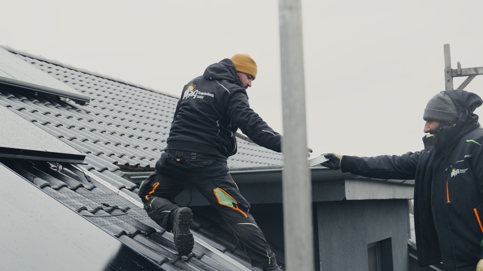 Zwei Monteure von RA Solartechnik bei der Arbeit auf einem Ziegeldach mit schwarzen Solarmodulen, einer reicht dem anderen Material an